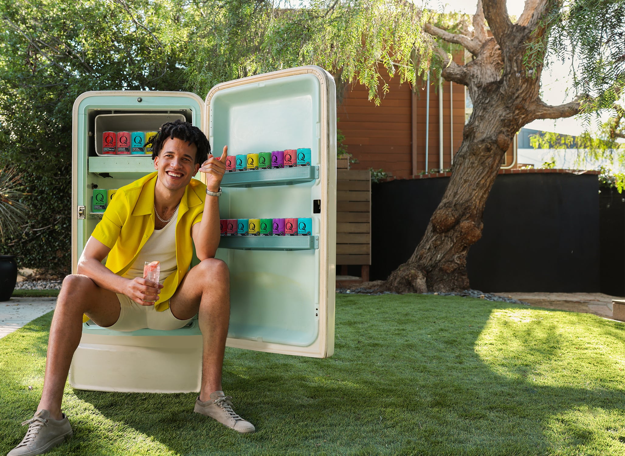A young man in Summer clothing sits by a fridge filled with Q Mixers cans in a sunny backyard